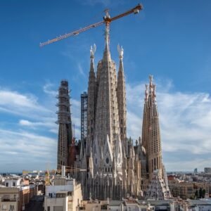 A large basilica under construction with several tall spires and a crane on top, set against a clear blue sky.