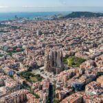 Vista aérea de una ciudad densamente poblada con una gran y detallada catedral en el centro y una línea costera con el océano al fondo.