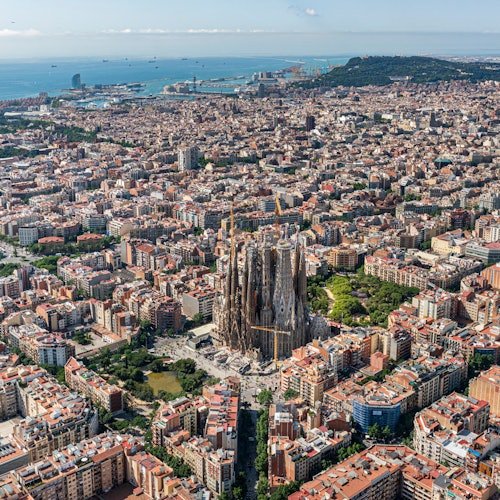 Vista aérea de una ciudad densamente poblada con una gran y detallada catedral en el centro y una línea costera con el océano al fondo.