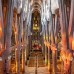 Interior de una gran catedral con altas columnas ornamentadas, vibrante iluminación de vidrieras y disposición simétrica de los asientos.
