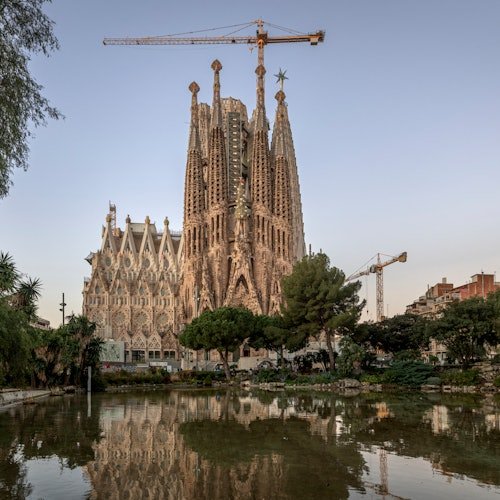 La basílica de la Sagrada Familia de Barcelona, con grúas sobre ella, reflejada en un tranquilo estanque rodeado de árboles.