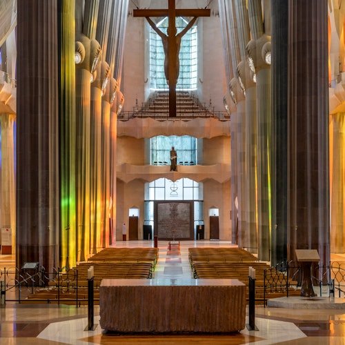Interior de una catedral con altas columnas, vidrieras y un crucifijo colgado sobre el altar. Los bancos están alineados en filas.