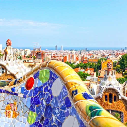 Colorful mosaic bench overlooking a cityscape with unique architectural buildings and distant skyline under a clear blue sky.