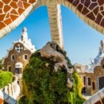 Un arco de mosaico de piedra enmarca caprichosos edificios con detalles arquitectónicos únicos, vegetación y un cielo azul despejado de fondo.