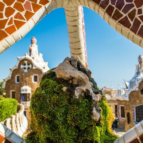 Un arco de mosaico de piedra enmarca caprichosos edificios con detalles arquitectónicos únicos, vegetación y un cielo azul despejado de fondo.