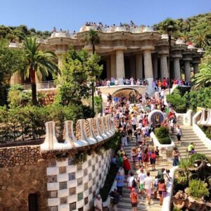 A bustling crowd explores Gaudí's Park Güell, with iconic architecture, lush greenery, and a sunny blue sky overhead.