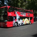 Autobús rojo de dos pisos con la marca "Madrid City Tour" e ilustraciones de dibujos animados de colores en el lateral, aparcado en una calle.