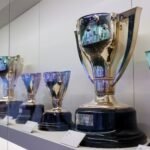 Several trophies of varying sizes are displayed on shelves, each with a plaque and card, inside a glass case.