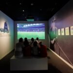 A group of people watching a soccer match on a screen in a dim room with images and text on the side walls.