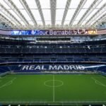 Interior view of a large stadium with green field, blue seating, and "REAL MADRID" displayed in white letters on the stands.
