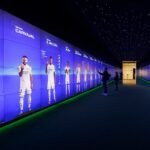 A futuristic display wall showing life-sized images of soccer players in a dimly lit room with visitors interacting.