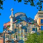 Un edificio colorido y ornamentado, con tejado ondulado, tejas decorativas y formas singulares, enmarcado por verdes árboles bajo un cielo azul.